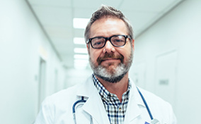 A male doctor smiles at the camera while standing in a hospital hallway.