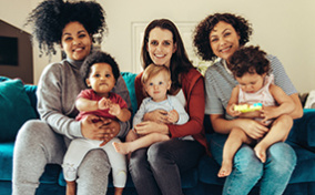 Three mons sit on a couch with babies on their laps. 