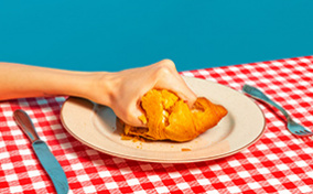 A hand crushes a croissant that sits on a plate on a red gingham table cloth.