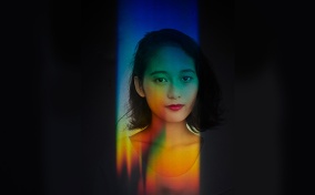 A woman looks at the camera with rainbow light all over her and black covering the periphery.