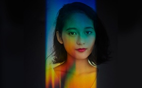 A woman looks at the camera with rainbow light all over her and black covering the periphery.