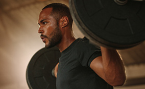 A man holds a bar bell on his shoulders and looks ahead with a concentrated expression. 