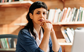 A nonbinary person sits with their elbows on a desk and looks at the camera.