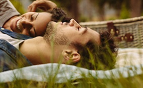 A man lies on a picnic blanket in the grass looking at the sky and a woman laughs on his chest.