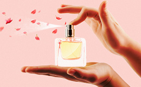 A pheromone perfume bottle rests in an open palm while the other hand sprays the perfume and rose petals. 
