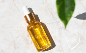 A dropper bottle of essential oil lays against a textured surface with green leaves casing a shadow nearby.