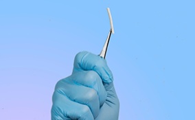 A hand inside of a surgical glove holds a birth control implant between a pair of tweezers.