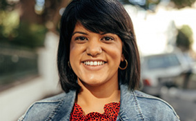 A woman in her 30s with shoulder length hair and bangs smiles at the camera on a sidewalk. 