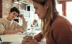 A man looks over at his partner while both are sitting at a table together on their laptops.