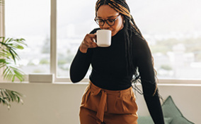 A woman stands at her desk drinking coffee from a mug and looking down at her laptop.