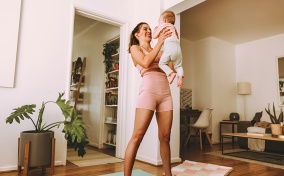 A woman in workout attire stands on a yoga mat while holding up her baby.