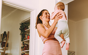 A woman in workout attire stands on a yoga mat while holding up her baby.