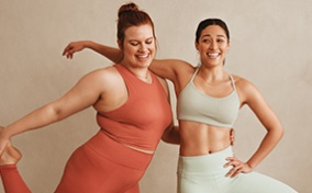 Two women stretch side-by-side in workout attire with their arms wrapped around each other.