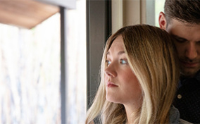 A couple stand by the window with a man hugging a woman from behind.