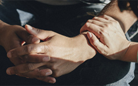 Two hands are folded together with another reaching out to hold his arm on the wrist.