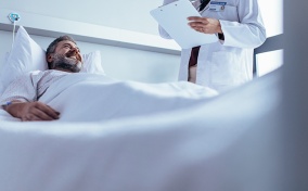 A man smiles in a hospital bed while looking up at his doctor after a vasectomy.