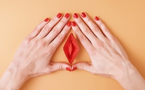Two hands with red painted nails surround frame a vulva-shaped origami. 