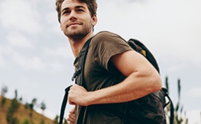 A young man wearing a backpack looks over his shoulder, smiling, with a mountain in the background.