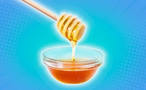 Honey is falling down from a dipper into a small dish against a blue background.