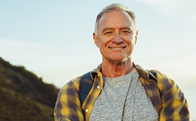 An older man is standing on a mountain and smiling at the camera with his arms folded.