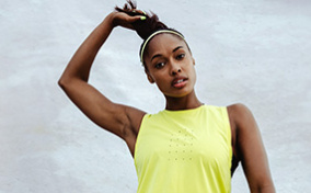 A woman in a yellow sleeveless shirt reaches to tug upwards on her ponytail.