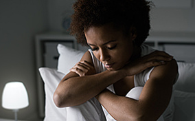 A grieving woman sits in bed with her legs up and arms folded over her knees.