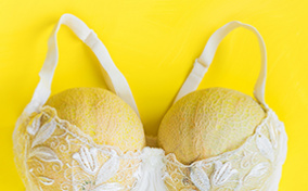 Two honeydew melons fill out a white, lacy bra on a yellow background.