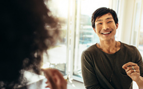 A man smiles at his friends in the kitchen as he brings food to his mouth. 