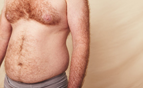 A shirtless man with a hairy chest and some belly fat stands in front of a beige background.
