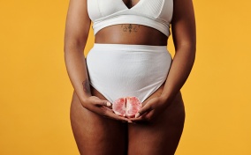 A woman in a white bra and high waisted underwear holds an open grapefruit in front of her crotch.