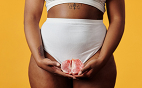 A woman in a white bra and high waisted underwear holds an open grapefruit in front of her crotch.