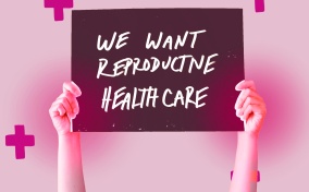 A sign is held up that says We Want Reproductive Healthcare with pink crosses around it.