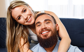 A smiling woman wraps her arms around a smiling, bearded man. 