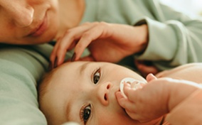 A mother touches her baby's head while it holds its pacifier and looks at the camera.