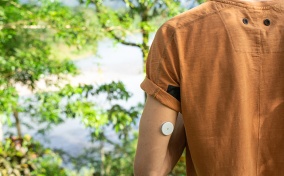 A man with diabetes stands near trees with his back to the camera with a glucose meter on his arm.