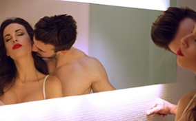A man and woman embrace in front of a mirror while he kisses her neck.