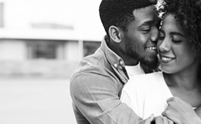 A smiling man wraps his arm around a smiling woman from behind.