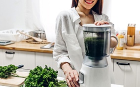A smiling woman in a kitchen stands next to a cutting board with parsley on it and places her hands on a blender.