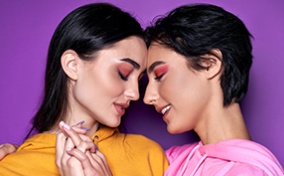 Two people with pink eye shadow press their foreheads together and hold hands in front of a purple background.