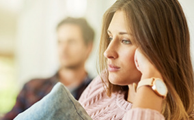 A woman stares blankly ahead with her head resting on her fist and her male partner sitting in the background.
