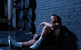 A bearded, shirtless man sits on the floor of a gym with his back against the wall. 