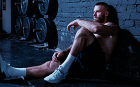A bearded, shirtless man sits on the floor of a gym with his back against the wall. 