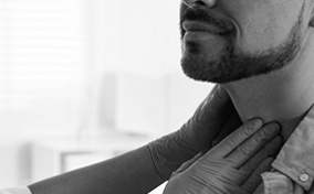 Two gloved hands palpate a bearded man's neck.