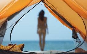 A nude woman standing near the ocean is out of focus and seen through the opening of a tent.