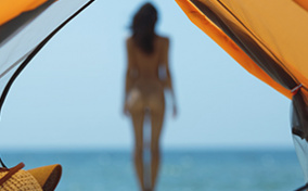 A nude woman standing near the ocean is out of focus and seen through the opening of a tent.