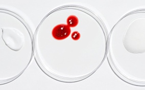 A petri dish with drops of period blood sits between two other petri dishes with skin care products.
