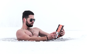 A man is doing cold water therapy outside in the opening of a snowy iced-over lake while reading a book.