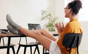 A woman sits with her legs propped up on a table and a cup in hand during a video call on her laptop.