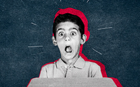 A little boy sits at a desk and makes a fearful expression with his eyes and mouth wide open.