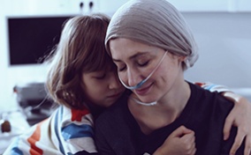 A young child hugs their mother who is wearing a head scarf and oxygen cannula.